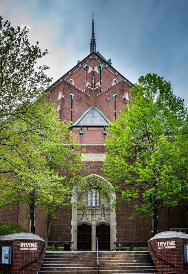 Exterior shot of Irvine Auditorium showing its red brick facade and church-like steeple. The building is framed by two lush green trees.